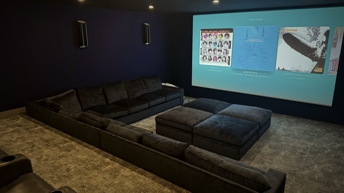 Restrepo Innovations home theater install with sectional, ottomans, and lit projection screen showing album art